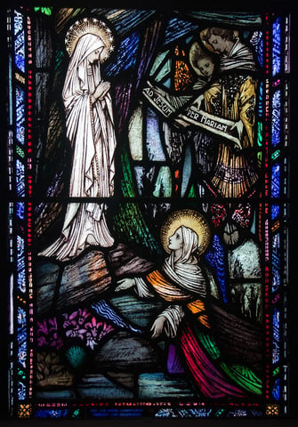 Stained glass depiction of St. Bernadette kneeling and looking up at the Blessed Virgin during the apparition at Lourdes.