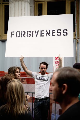 man on crowded street wears sorry shirt and holds forgiveness sign